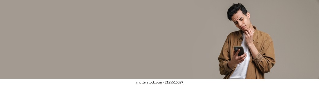 Pensive frowning young brunette man in t-shirt standing over gray wall background - Shutterstock ID 2125515029