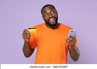 Pensive funny young african american man 20s wearing basic casual orange t-shirt standing using mobile cell phone hold credit bank card isolated on pastel violet colour background, studio portrait