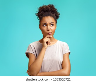 Pensive girl being deep in thoughts, raises eyebrows, curves lips, holds chin. Photo of african american girl wears casual outfit on turquoise background. Emotions and pleasant feelings concept.