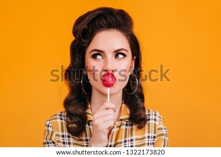 Pensive girl with elegant hairstyle licking candy. Studio shot of pinup woman with lollipop isolated on yellow background.