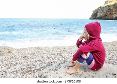 Pensive Girl Relaxing On A Pebbly Beach, Eating And Observing The Sea After Swimming, Barefoot And Dressed In Leggings And Fleece. Family And Children On Vacation, Late Summer Concept. 