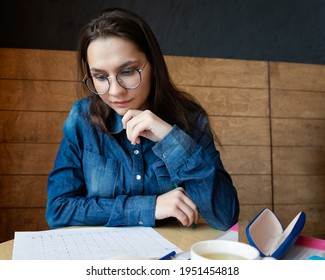 A Pensive Girl In Round Glasses Sits In A Cafe And Looking At The Drawing, She Fills Out A Lifestyle Schedule Drawn By Her With Felt Tip Pens, Portrait, A Business Photo Session