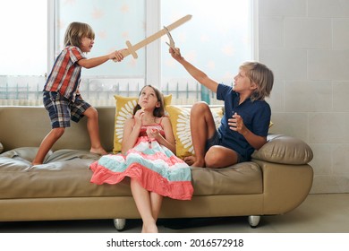 Pensive girl sitting on sofa and eating potato chips when her brothers playing with cardboard swords