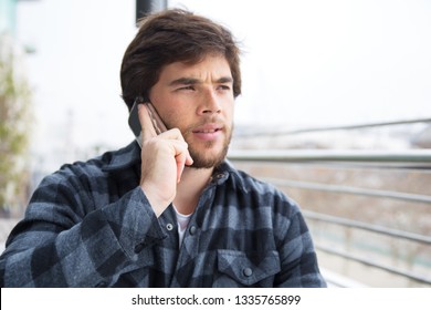 Pensive guy talking on cell on terrace. Serious young man in casual speaking on mobile phone outdoors and staring into distance. Phone talk concept