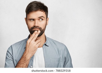 Pensive handsome male model keeps hand on chin, looks thoghtfully aside, thinks about something very important, tries to make decision, isolated over white background with copy space for your text