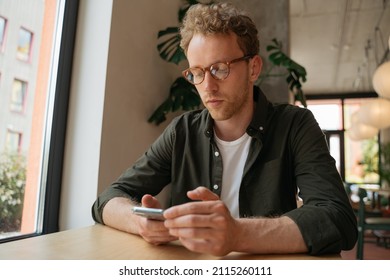 Pensive handsome man wearing eyeglasses using mobile phone, communication searching online sitting in modern cafe. Stylish hipster holding smartphone with mobile, online shopping, ordering something 