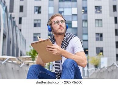 A Pensive Handsome Young Man In A White T-shirt, Wearing Headphones, Sits On The Steps With A Notebook, Diary Or Sketchbook, In A City Park. Life Sketch.