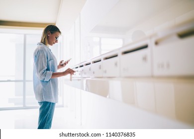 Pensive Hipster Girl In Casual Wear Using Key For Unlock Mailbox In Apartment Hall, Woman Checking Notification On Mobile Phone Open Metal Letterbox For Get Correspondents From Postal Service