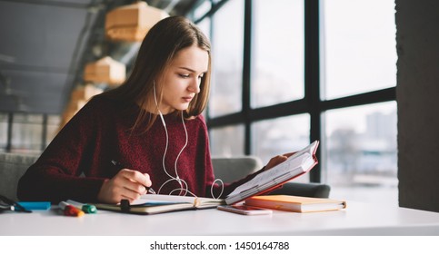 Pensive hipster girl learning language online via earphones using application while writing new words into textbook, attractive smart woman enjoying playlist music at cafeteria and studying