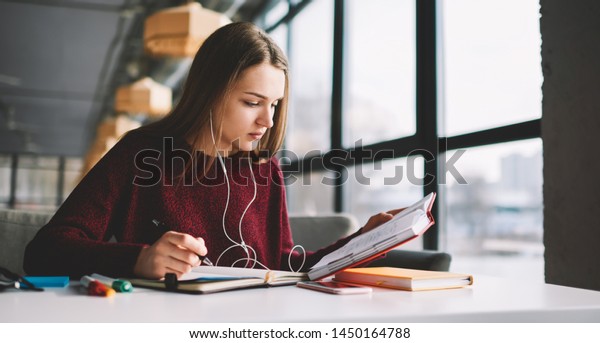 Pensive hipster girl learning language online\
via earphones using application while writing new words into\
textbook, attractive smart woman enjoying playlist music at\
cafeteria and studying