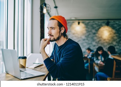 Pensive hipster guy in earphone looking away listening music via app on laptop computer, dreamy male caucasian student enjoying audiobook thinking on ideas for blog publication in coworking space