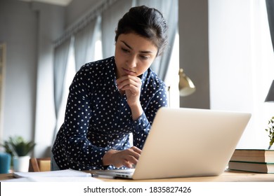 Pensive Indian Business Woman Standing In Office Room Standing Lean Over Workplace Desk Working On Laptop Thinking About Problem Solution, Create Research, Do Paperwork, Corporate Task Prepare Report