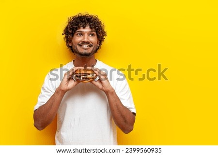 pensive indian guy eating a delicious burger on a yellow isolated background and thinking, young indian man holding fast food and dreaming and imagining