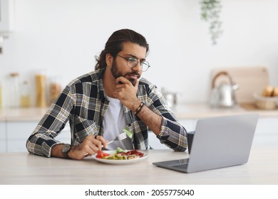 Pensive Indian Man Freelancer Having Snack While Working From Home During Coronavirus Pandemic, Stressed Arab Guy Eating Healthy Food, Looking At Laptop Screen, Need For Vacation, Copy Space