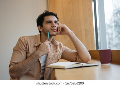 Pensive Indian Man Working, Planning Start Up, Thinking, Holding Pen Near Face, Sitting At Workplace. Busy Student Studying, Exam Preparation. Portrait Of Mixed Race Writer Brainstorming 