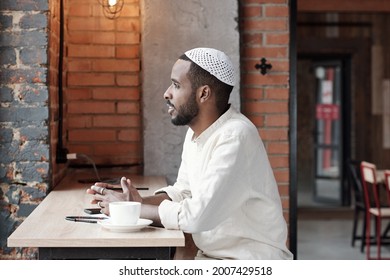 Pensive Inspired Young Islamic Man In Kufi Cap Sitting At Table In Loft Cafe And Looking Out Window