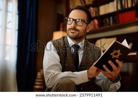 Pensive Jewish man enjoying in reading Hebrew bible at home and looking at away.