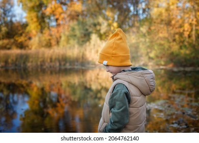 Pensive Little Boy Stands On The Bank Of River, Admires The Magnificent View Of Autumn Forest And Its Reflection In The Lake Water. Autumn Walk. Outdoor Lifestyle, Active Family Lifestyle