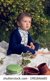 Pensive Little Boy Of Two Years Old In A Nice Suit Is Sitting On White Carpet With Blooming Brunches On It And Blooming Shrubs In The Background. Far Away Look Of Thoughtful Child.