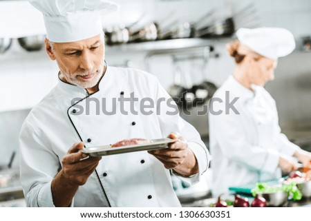 pensive male chef in uniform holding meat dish on plate with colleague on background in restaurant kitchen