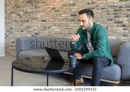 Pensive male professional holding eyeglasses and looking at laptop while sitting on sofa in office