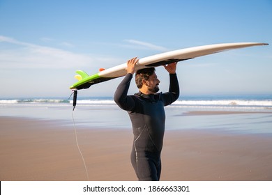 Pensive Male Surfer In Wetsuit Walking On Beach And Carrying Surfboard Overhead. Medium Shot. Surfing And Active Lifestyle Concept