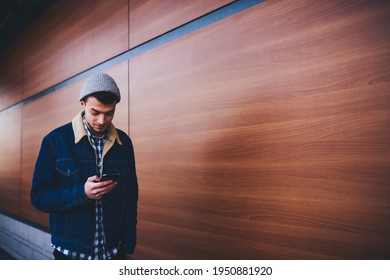 Pensive male in trendy clothes walking on street near building with wooden wall and surfing mobile phone having free time