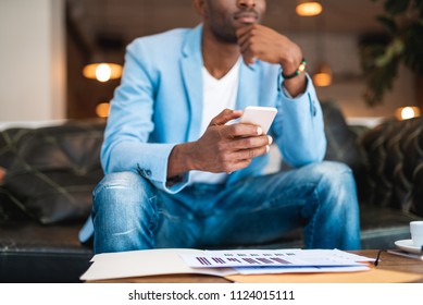 Pensive Male Typing In Modern Phone While Locating On Cozy Couch Opposite Table With Graphics