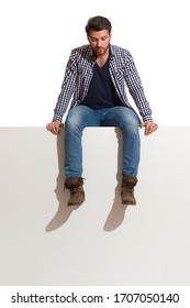 Pensive Man In Boots, Jeans And Lumberjack Shirt Is Sitting On A Top And Looking Down. Full Length Studio Shot Isolated On White.