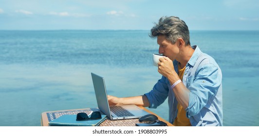 Pensive Man Freelancer Working On Laptop Computer At Beach By Blue Sea, Drinking Coffee. Concept Remote Work, Freelance