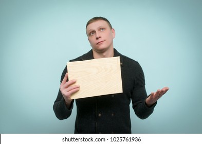 Pensive Man Is Holding In Front Of Him A Wooden Board With Copy Space And Is Thinking Isolated On Blue Background. Obvious Choice Concept.