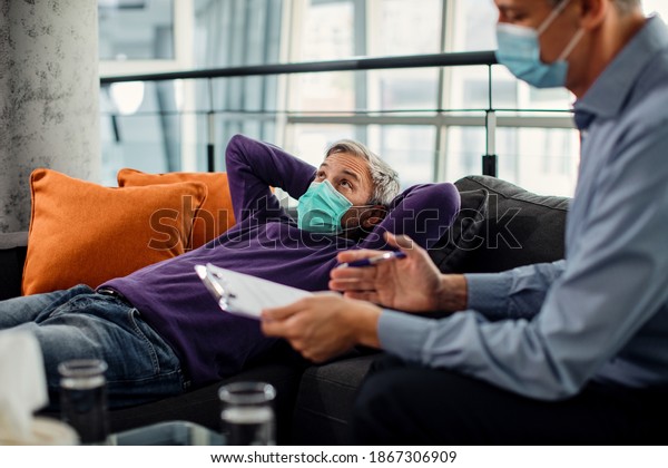 Pensive man lying down on\
psychiatrist\'s couch and talking to his therapist during\
coronavirus pandemic. 