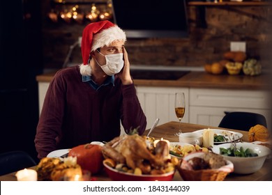 Pensive Man Missing His Family While Sitting Alone At Dining Table And Wearing Face Mask Due To Coronavirus Epidemic. 