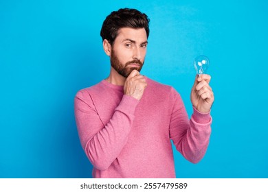 Pensive man in pink sweater contemplates idea with light bulb against bright blue background - Powered by Shutterstock