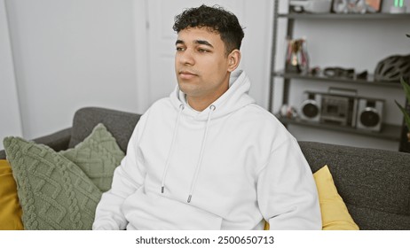 A pensive man wearing a hoodie sits casually in a modern living room with stylish decor. - Powered by Shutterstock