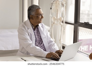 Pensive mature 65s Indian therapist in white lab coat working on his laptop seated at workplace desk, prescribe medications remotely, check current health status of patient via telemedicine platform - Powered by Shutterstock