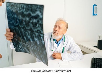 Pensive Mature Adult Male Doctor Looking MRI Spine Image Of Patient Sitting At Desk With Computer In Medical Office Room, Thinking About Diagnosis, Expressing Concern Of Illness.