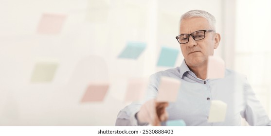 Pensive Mature Businessman In Glasses Putting Sticky Notes On Window At Office, Empty Space - Powered by Shutterstock