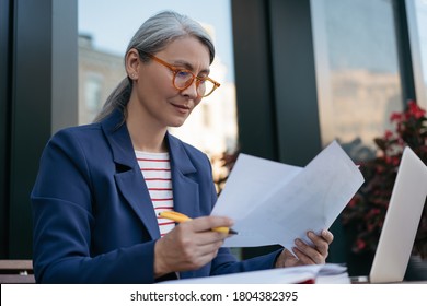 Pensive mature businesswoman reading contract, planning project, brainstorming. Portrait of middle aged asian secretary reading, working with documents, sitting at workplace  - Powered by Shutterstock