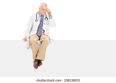 Pensive Mature Doctor Sitting On A Blank Panel Isolated On White Background