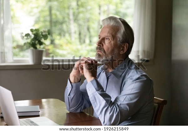 Pensive mature grey haired 60s - 70s aged man\
sitting at table with laptop at home, looking away lost focus.\
Thoughtful retired OAP working at computer from home, thinking over\
retirement during break
