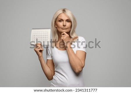 Pensive Mature Lady Holding Blank Menstrual Calendar And Touching Chin, Thoughtful Senior Female Suffering Lack Of Menstruation, Having Climacteric Symptoms Or Menopause, Standing On Grey Background