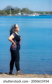 Pensive Mature Latin Woman Standing In Profile On The Shore Of Lake Grevelingen, Sailboats Anchored In Blurred Background, Black Clothes, Tanned Skin, Sunny Day In Zonnemaire, Zeeland, Netherlands