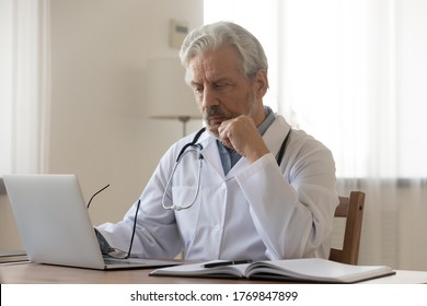 Pensive Mature Male Doctor Or GP Sit At Desk In Hospital Look At Laptop Screen Thinking Of Patient Diagnosis, Thoughtful Old Man Physician Or Therapist Work On Computer In Cabinet, Make Decision