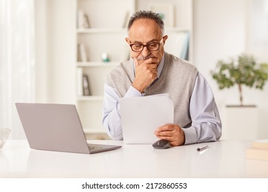 Pensive Mature Man Sitting In Front Of A Laptop Computer And Looking At A Paper Document