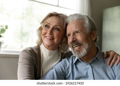 Pensive Mature Retired Family Couple Looking Away At Window And Smiling. Happy Healthy Married Pensioners Thinking Of Carefree Retirement, Planning Future Vacation And Leisure Time, Hugging Together