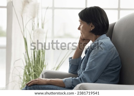 Pensive mature woman sits on armchair staring into distance, deep in thoughts, pondering, thinks on life problems, spend time alone at home, suffering from solitude and boredom on retirement. Concerns