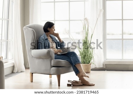 Pensive mature woman working on laptop at home, sitting on armchair with computer on laps, staring into distance, thinking, search problem solution, ponder bad on-line news feels concerned or fatigued