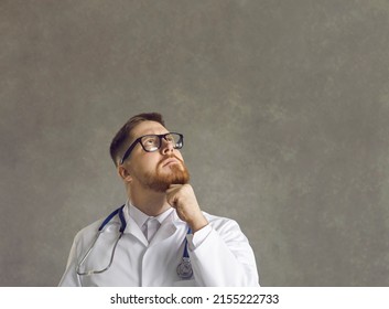 Pensive Medical Doctor Thinking On Question About Health On Studio Headshot Portrait. Thoughtful Caucasian Male Practitioner In Uniform Looking Up With Hand On Chin Making Clinical Decision