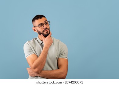 Pensive Middle Eastern Guy Thinking And Touching Chin, Looking Aside At Free Space, Posing Over Blue Background, Studio Shot. Let Me Think Concept